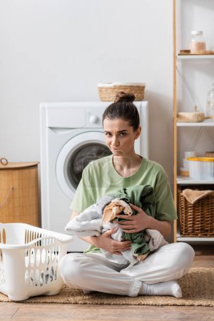 Téléchargez les photos : Jeune femme perplexe tenant des vêtements près du panier et machine à laver dans la buanderie - en image libre de droit