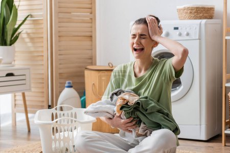 upset woman holding clothes near basket and crying in laundry room 