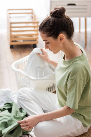 Téléchargez les photos : Vue latérale de la femme triant les vêtements près du panier pendant les travaux ménagers - en image libre de droit