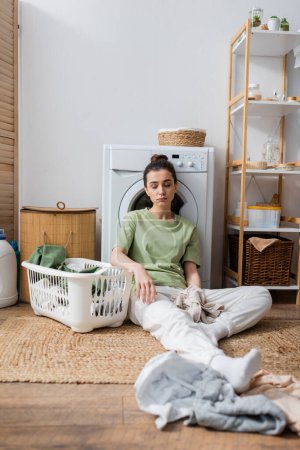 Foto de Mujer cansada sentada cerca de la ropa y la lavadora en la lavandería - Imagen libre de derechos
