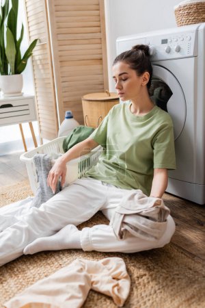 Tired woman holding clothes near basket and washing machine at home 