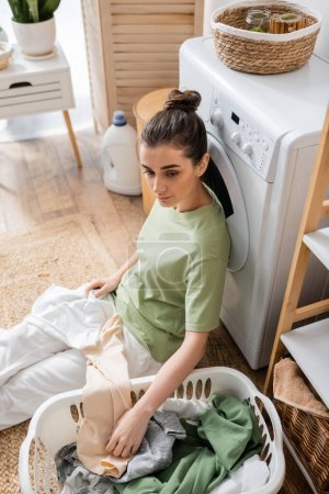 Vue grand angle de la femme mécontente assise près du panier avec des vêtements et une machine à laver à la maison 