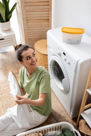 Vue grand angle de la femme souriante regardant la boîte sur la machine à laver près du panier avec des vêtements dans la buanderie 