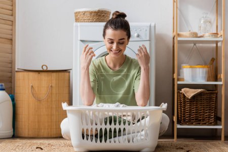 Positive brunette woman looking at basket with clothes in laundry room 