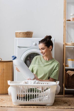 Fröhliche brünette Frau hält Waschmittel neben Korb mit Kleidung in der Waschküche 