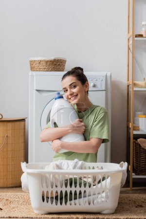 Positive Frau hält Waschmittel in der Hand und blickt in Waschküche in die Kamera 