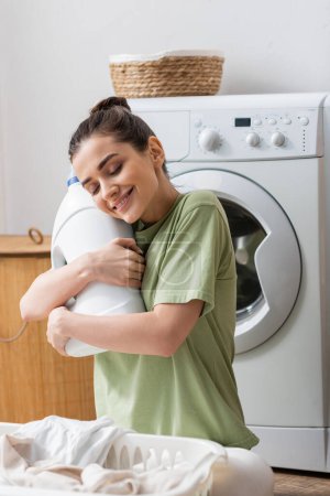 Cheerful young woman hugging bottle with washing liquid near basket with clothes in laundry room 