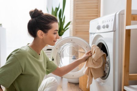 Foto de Side view of young woman putting clothes in washing machine at home - Imagen libre de derechos