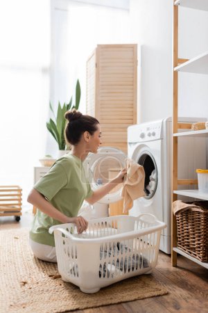 Foto de Side view of smiling woman putting clothes in washing machine at home - Imagen libre de derechos