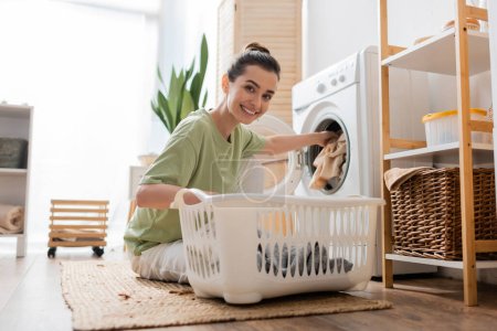 Jeune femme souriant à la caméra tout en mettant des vêtements dans la machine à laver dans la buanderie 