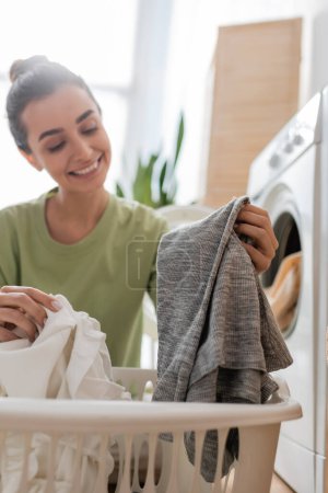 Blurred young woman holding clothes near basket and washing machine at home 