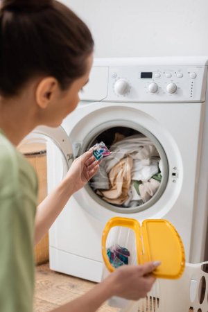 Mujer joven borrosa sosteniendo cápsula de lavado cerca de la máquina en la sala de lavandería 