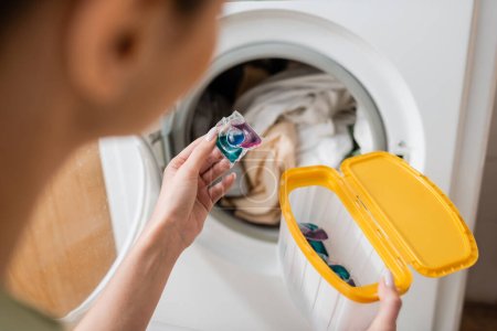 Vista recortada de una mujer borrosa sosteniendo la cápsula de lavado y la caja cerca de la máquina en la sala de lavandería 