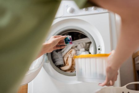 Ausgeschnittene Ansicht einer Frau, die Flüssigwaschmittel in der Nähe von Kleidung in der Waschmaschine zu Hause hält 