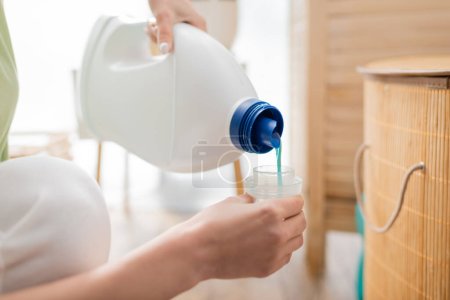 Cropped view of woman pouring washing liquid in bottle cap in laundry room 