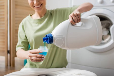 Ausgeschnittene Ansicht einer lächelnden Frau, die Flüssigwaschmaschine in Waschküche gießt 
