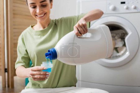Mujer sonriente borrosa vertiendo líquido de lavado en la tapa cerca de la máquina en la sala de lavandería 