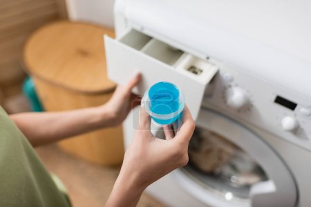 Cropped view of woman holding cap with blue washing liquid near machine in laundry room 