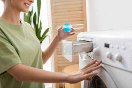 Vista recortada de una mujer sonriente sosteniendo un limpiador líquido cerca de la lavadora en casa 