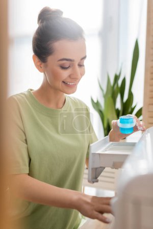 Lächelnde Frau hält Waschmittel neben Maschine in Waschküche 