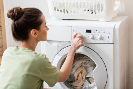 Téléchargez les photos : Vue latérale de la femme floue commutant la machine à laver près du panier dans la buanderie - en image libre de droit