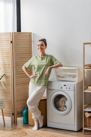 Smiling woman looking at camera while standing near washing machine in laundry room 