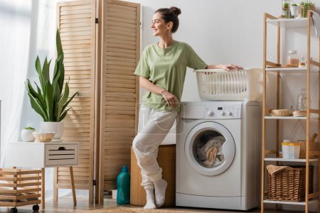 Smiling woman standing near basket and washing machine at home 