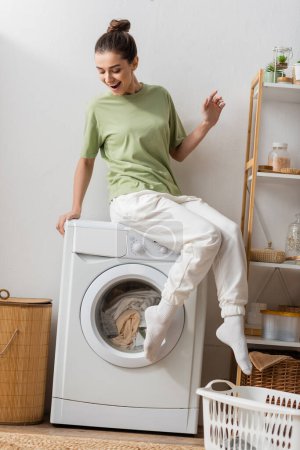 Aufgeregte junge Frau sitzt auf Waschmaschine in Waschküche 