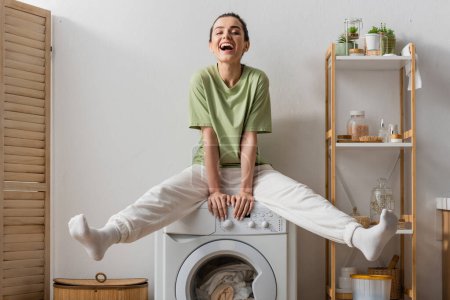 Excited woman sitting on washing machine with clothes at home 