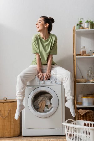 Aufgeregte junge Frau sitzt auf Waschmaschine in Waschküche 