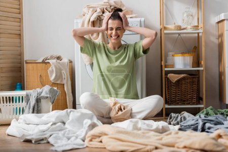 stressed young woman sitting near clothes on floor in laundry room 