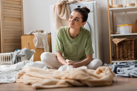Displeased woman sitting near blurred clothes in laundry room 