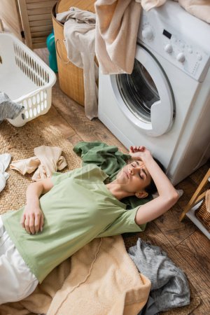Top view of tired woman lying around clothes near washing machine in laundry room 