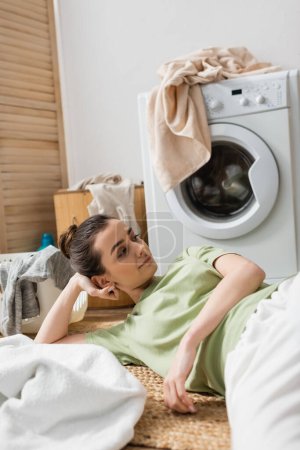 Brunette woman lying near clothes on floor in laundry room 