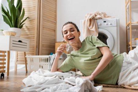Fröhliche Frau liegt neben Kleidung und Korb in Waschküche auf dem Boden 