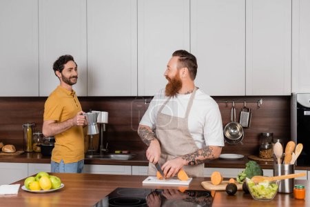 Smiling gay man holding coffee and cups near husband cooking in kitchen 