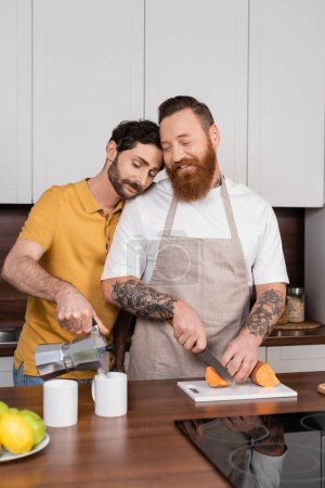Gay man pouring coffee near smiling husband cutting sweet potato in kitchen 