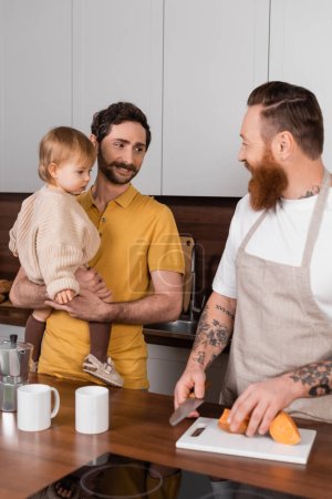 Smiling gay father holding daughter while husband cooking in kitchen 