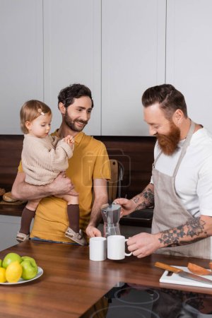 Smiling gay man pouring coffee near partner with daughter in kitchen 