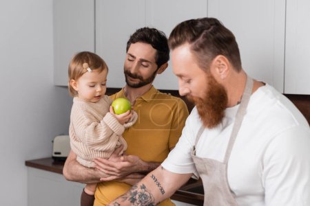 Smiling gay man holding daughter with apple near bearded partner in kitchen 