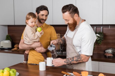 Bearded gay man pouring coffee near husband holding toddler daughter in kitchen 