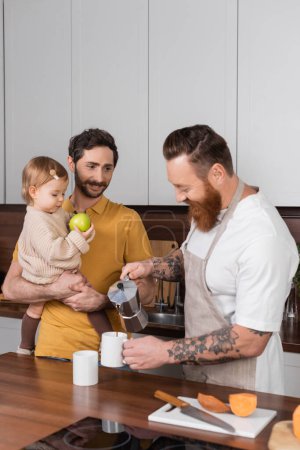 Smiling gay man holding toddler daughter with apple while partner pouring coffee in kitchen 