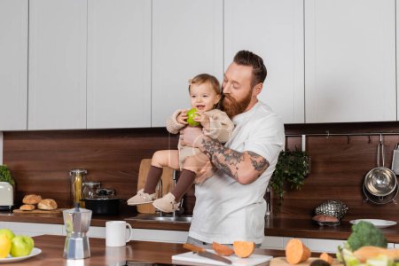 Tattooed dad holding daughter with apple in kitchen 