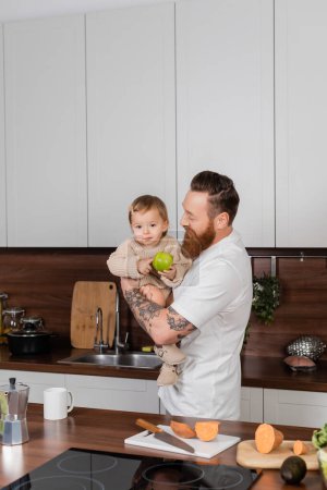 Bearded man holding toddler daughter with apple in kitchen at home 