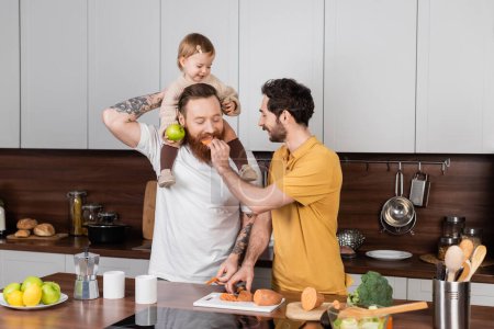 Gay man feeding partner near baby daughter in kitchen 