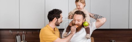 Gay man feeding husband near smiling daughter with green apple in kitchen, banner