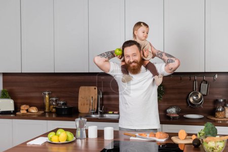 Smiling tattooed man holding toddler daughter with apple near vegetables in kitchen 