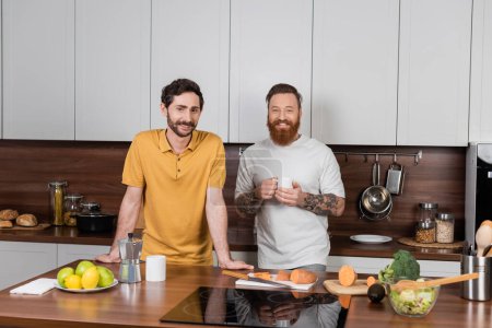 Smiling gay couple with coffee looking at camera in modern kitchen 