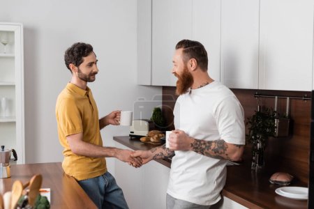 Foto de Gay couple with cups of coffee holding hands in kitchen at home - Imagen libre de derechos