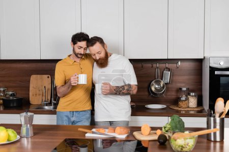 Gay couple holding cups of coffee near vegetables in kitchen 